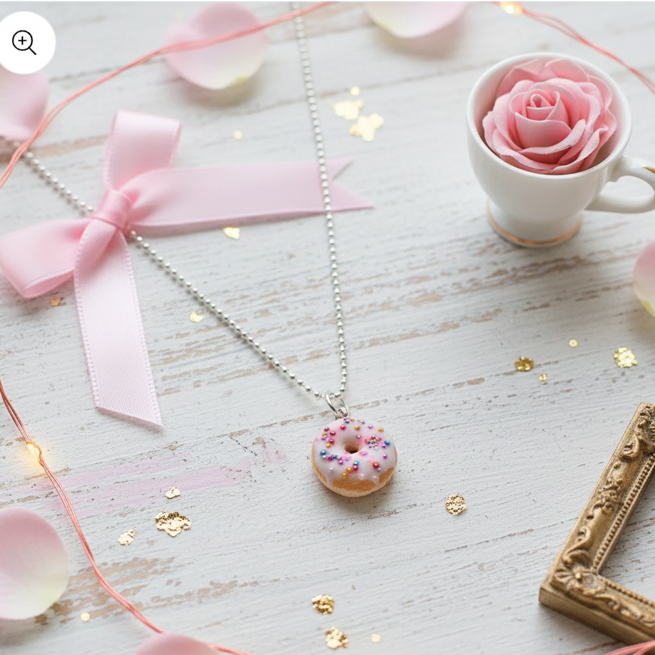 Necklace with a funfetti donut pendant on a decorative surface with a cup of pink flowers, pink ribbon, and gold frame.