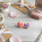 Heart-shaped  sugar cookie earrings on a wooden surface with a teacup, gift box, and fork in the background.